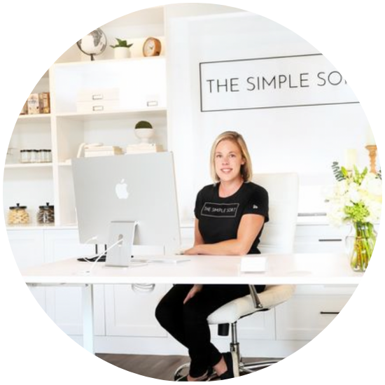 A woman sitting confidently at a home office desk with a large Apple computer, wearing a black t-shirt with 'The Simple Sort' logo, surrounded by stylish home decor and flowers.